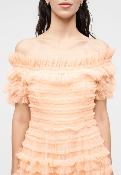 Naine kannab virsikuvärvi õlgadeta kleiti mitme kihilise paelade ja lühikeste puhvervarrukatega, tavalise tausta ees.