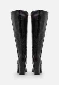 Bottes noires montant jusqu'au genou en cuir embossé crocodile, avec un talon épais et une fermeture éclair latérale pour un accès facile.