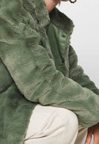 Cappotto in pelliccia ecologica verde con una texture morbida, caratterizzato da una chiusura a bottoni a pressione e una fodera verde liscia, indossato sopra un top di colore chiaro.