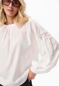 Kobieta w dużych okrągłych okularach w stylu żółwi, białej bluzce z koronkowymi wycięciami na puffowanych rękawach oraz czarnych spodniach w prążki.