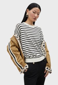 Gestreifter Pullover in Creme und Marine, kombiniert mit einer beige Jacke mit Punkten. Schwarze Hosen ergänzen die Farbpalette des Outfits.