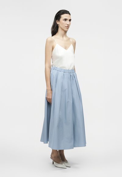 Γυναίκα με μακριά σκούρα μαλλιά που φοράει λευκό αμάνικο τοπ, φούστα μέσης ψηλής σε ανοιχτό μπλε midi μήκος και λευκά μυτερά τακούνια, στέκεται στραμμένη προς τα αριστερά.