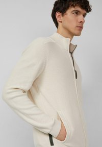 Lichtbeige trui met ritssluiting, voorzien van een geribde kraag en manchetten. Heeft zijzakken met donkere accenten. Zachte, gestructureerde stof.