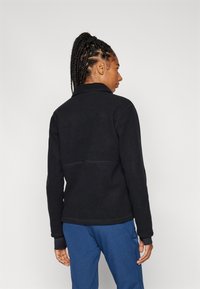Čierna fleece bunda s vysokým golierom, s hladkou textúrou a kontrastným prešívaním. V páre so modrými nohavicami, zobrazená zozadu.