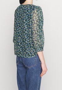 Květinová blůza v tmavě zelené barvě s modrými a žlutými květy, průhledné pouzdrové rukávy, elastické manžety a kulatý výstřih, spárovaná s džínami s vysokým pasem.