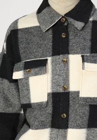 Cămașă flanel cu carouri alb-negru, cu două buzunare pe piept, nasturi maroon și guler cu buton. Textură moale și croială lejeră.