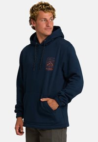 Młody mężczyzna ubrany w granatowy hoodie z przednią kieszenią i brązową grafiką na piersi, patrzący na bok na jednolitym tle.