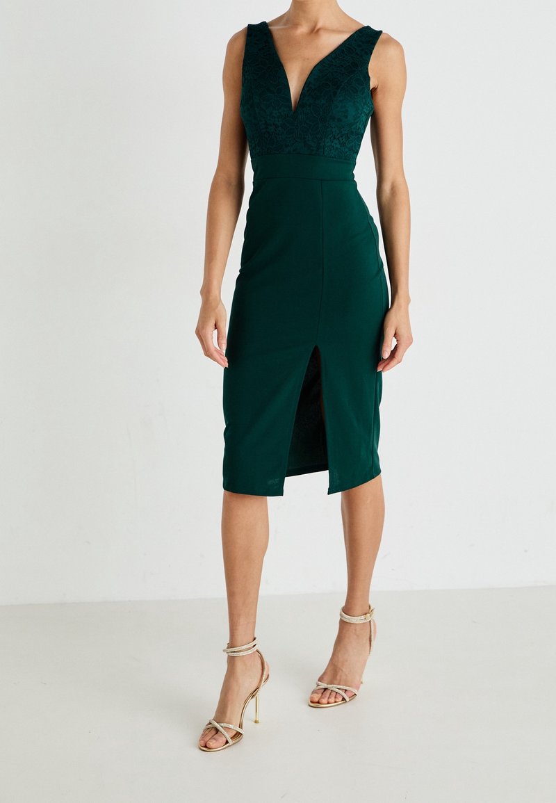 Smaragdfarbendes Kleid mit V-Ausschnitt, Spitzenoberteil, figurbetontem Schnitt, knielang und einem Schlitz vorne. Kombiniert mit goldenen Riemchen-High Heels.