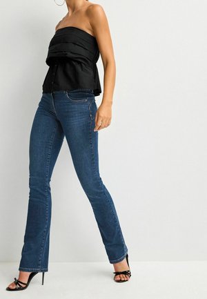 Femme portant un jean slim bleu foncé, un top bustier noir et des sandales à talons hauts noires sur fond blanc.