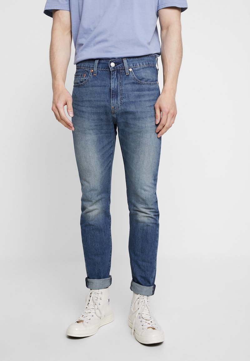 Blå denimjeans med en smal passform, ljusblekning, femficksdesign och knappstängning. I kombination med vita högstövlar.