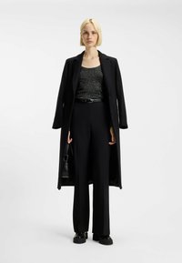 Donna con capelli biondi corti, vestita con un lungo cappotto nero, un top nero scintillante, pantaloni larghi neri e scarpe con piattaforma nere, mentre tiene una piccola borsa nera.