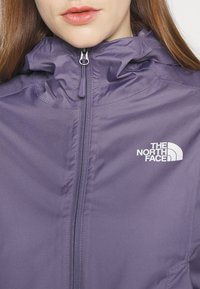 Chaqueta morada con cremallera, cuello alto, textura de tela lisa y el logo blanco de "The North Face" en el lado izquierdo.