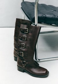 Hnědé kožené vysoké boty na kolena s několika kovovými přezkami a blokovým podpatkem. Design zahrnuje dekorační pásky a hladkou texturu.