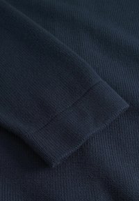 Marinblå stickad tröja ärm med ribbade mudd och strukturerad tyg. Närbild som visar det fina stickmönstret och den släta ytan.