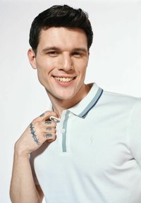 Biała koszulka polo z niebieskim i zielonym paskiem przy kołnierzyku, z dwoma guzikami z przodu oraz małym logo na piersi. Tatuaże widoczne na dłoni.