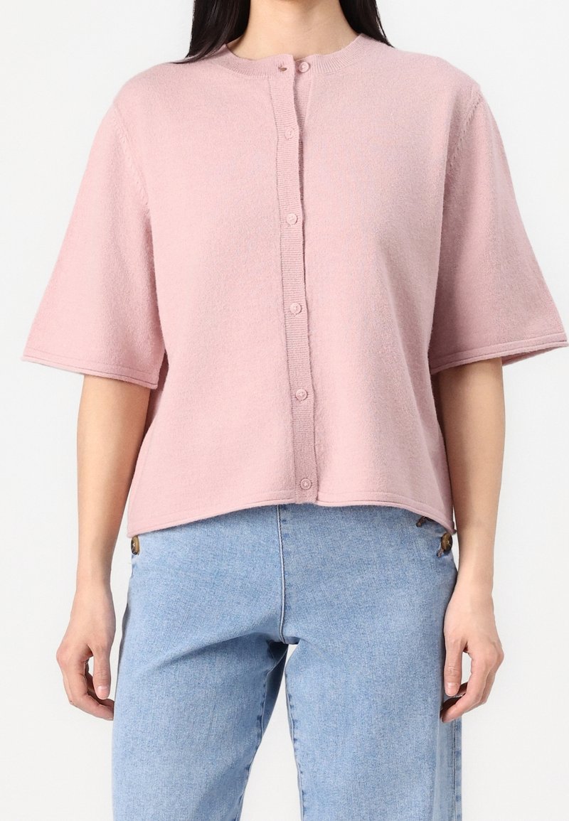 Femme portant un pull à manches courtes rose clair boutonné, associé à un jean taille haute bleu clair sur un fond uni.