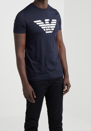 Homme portant un t-shirt bleu marine ajusté avec un grand logo blanc Emporio Armani et un pantalon noir, debout devant un fond uni.