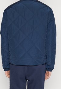 Tmavě modrá prošívaná bunda se vzorem diamantového prošívání, elastickými manžetami a hladkou texturou; má rovný střih a neformální design.