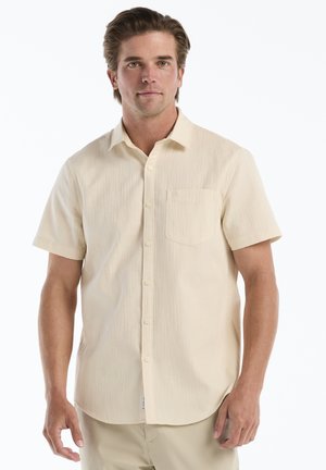 Mann trägt ein beige kurzärmeliges Hemd mit Brusttasche und steht vor einem einfarbigen weißen Hintergrund.