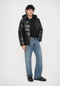 Junger Mann steht da und trägt eine schwarze, glänzende Pufferjacke, ein schwarzes Shirt, lockere blaue Jeans und schwarze Sneakers vor einem weißen Hintergrund.