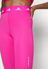 Różowe legginsy sportowe z szerokim pasem w talii, ozdobione trzema białymi, ukośnymi paskami. Materiał jest gładki, a logo znajduje się blisko kostki.
