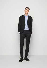 Juodas siūtas švarkas virš šviesiai mėlynos spalvos marškinių, derinamas su tamsiomis kelnėmis. Apranga pasižymi švariu dizainu ir klasikinio kirpimo. Juodi odiniai batai.