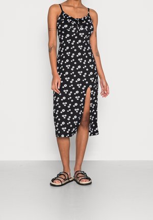 Black dress with white floral pattern, adjustable spaghetti straps, slit on one side, paired with black sandals. Smooth texture.