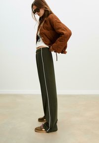 Hnedá prešívaná bomber bunda, biela grafická košeľa, tmavo zelené nohavice s bielymi bočnými pruhmi a hnedé slip-on topánky so šnúrkami.