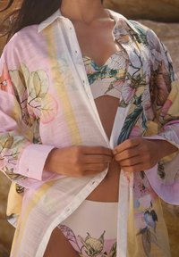 Camicia leggera e trasparente con bottoni in colori pastello e motivi floreali; indossata sopra un bikini stampato coordinato, mette in risalto un look estivo.