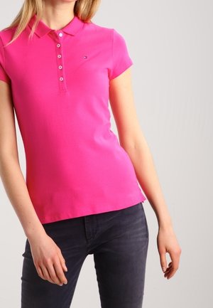 Kobieta ubrana w dopasowaną, jasnoróżową koszulkę polo z krótkim rękawem i guzikami oraz ciemne jeansy na jednolitym tle.