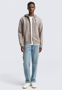 Mikina s kapucňou v svetlosivej farbe, vyrobená z mäkkej látky, s prednými vreckami a štýlovými šnúrkami. Kombinovaná so svetlomodrými džínsami a bielymi teniskami.