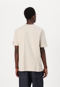 T-shirt beige à manches courtes en coton, avec un col rond et une coupe décontractée. Détail de couture notable près de l'épaule.