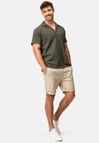 Kortærmet skjorte med knapper i olivengrøn, kombineret med opløftede beige shorts og hvide sneakers, der viser et afslappet, let design.