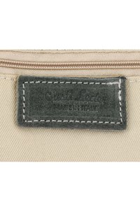 Étiquette en cuir gris avec couture blanche sur tissu beige, embossée d’un texte pâle « Made in Italy » sous un nom de marque.