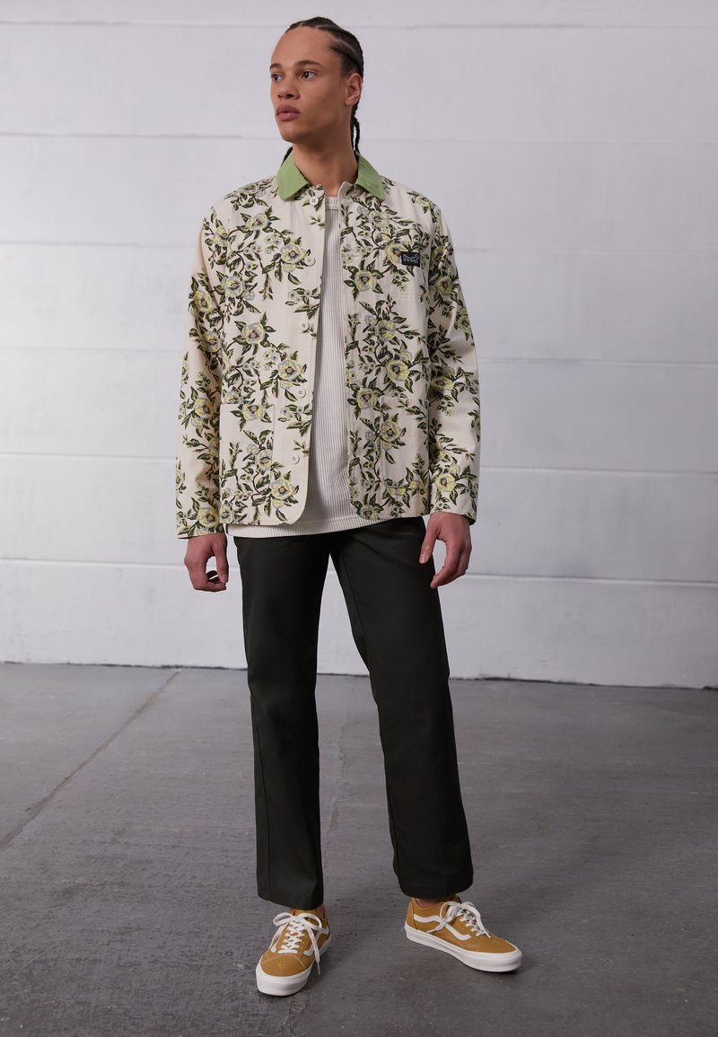 Floral patterned jacket in cream with green collar, worn over a textured off-white sweater, paired with dark olive pants and yellow sneakers.