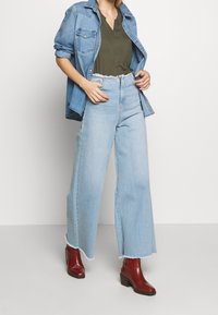 Lyseblå vide jeans med flosset kant, parret med en mørkegrøn v-hals top og en denim skjorte, suppleret med røde hælede ankelstøvler.