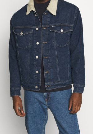 Mand iført en mørkeblå denimjakke med hvid fleece-krave, sort skjorte og mellemblå jeans, stående foran ensfarvet baggrund.