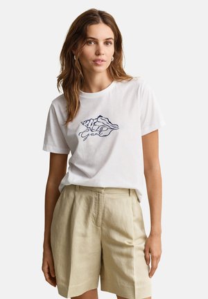 Femme aux cheveux châtain clair portant un t-shirt blanc à manches courtes avec un motif coquillage et un short beige taille haute, debout devant un fond uni.