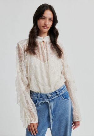 Cream lace blouse with frilled sheer sleeves, featuring a high collar and button closure, paired with light blue high-waisted denim jeans.