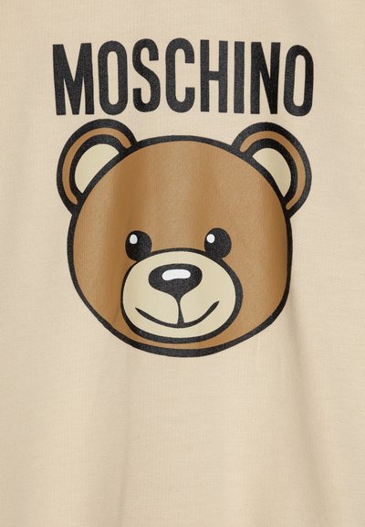 Beige bomull T-skjorte med et trykk av et tegneserie-bjørneansikt i brunt med sorte konturer, ledsaget av merkevarenavnet "Moschino" i fet sort tekst.