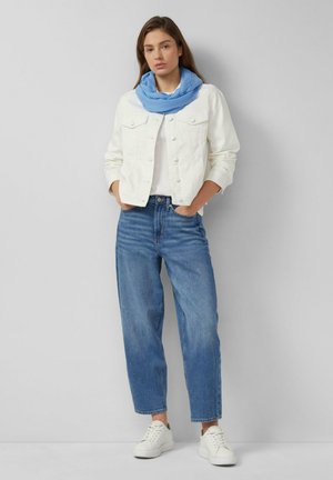 Woman standing with hands in pockets wearing white jacket, blue scarf, loose blue jeans, and white sneakers against a gray background.