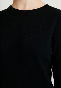 Gros plan d’une personne portant un pull noir en maille unie à col rond sur un fond clair.