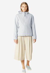 Lichtgrijze waterdichte jas met een capuchon, een rits aan de voorkant en zijzakken, gecombineerd met beige wijde broek en lichtblauwe geperforeerde instappers.