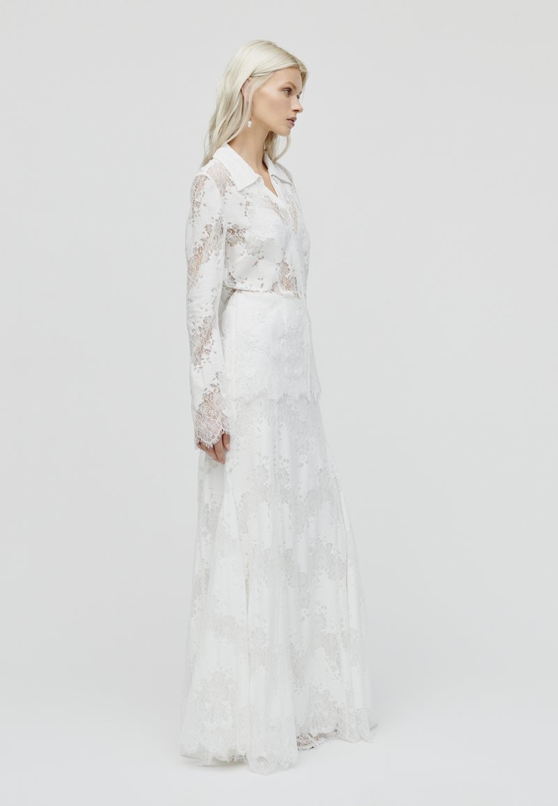 Weißes Spitzenkleid mit langen Ärmeln, ausgestattet mit einem spitzen Kragen und einem geschichteten Rockdesign, das kunstvolle Blumenmuster zeigt.