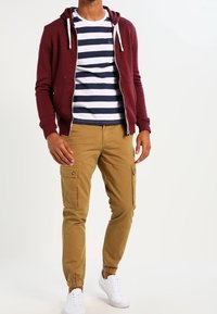 Körsbärsrandig zip-up hoodie med vita dragsnören, marinblå och vit randig t-shirt, ljusbruna cargobyxor med fickor, vita sneakers.