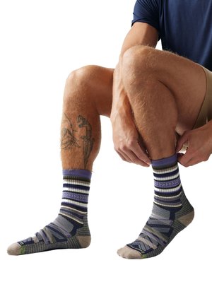 Jambes d'une personne assise portant des chaussettes rayées violettes et grises, ajustant une chaussette, avec un tatouage d'une dague sur le mollet gauche.