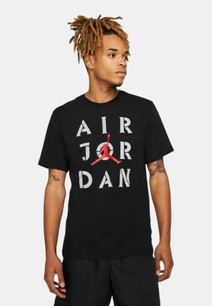 Joven con rastas que lleva una camiseta negra de Air Jordan con letras blancas y un logo rojo de Jumpman, mirando hacia su derecha.