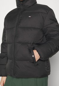 Giacca a vento nera con colletto alto, chiusura a zip e tasche laterali. Il tessuto ha una consistenza liscia con sezioni cucite per l'isolamento.