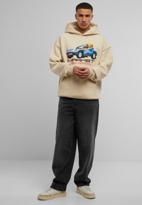 Beige hettegenser med grafikk av en blå bil og en bjørn, kombinert med mørkegrå vide jeans og beige joggesko med hvite detaljer.