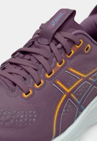 Vijolična športna čevlja z teksturiranim mrežastim zgornjim delom, ki ima oranžne in modre poudarke, s sistemom vezalke in oblazinjenim jezikom. Bel podplat.